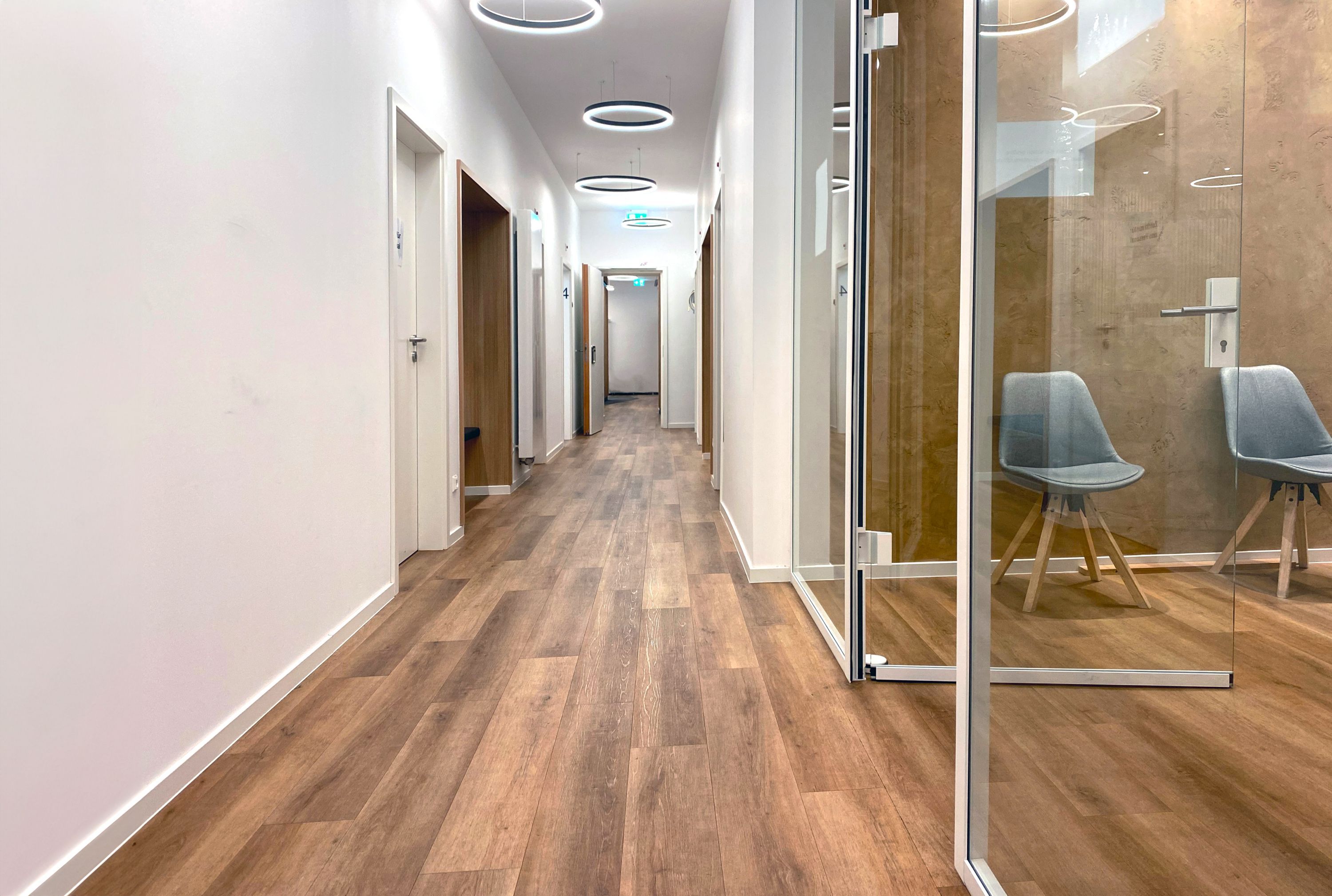 Flur mit Parkettboden, rechts einige Stühle hinter einer Glasscheibe